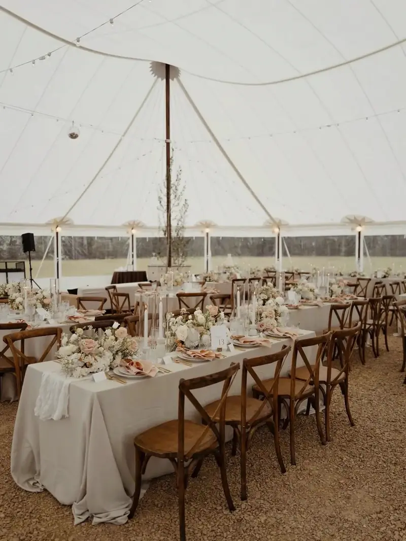 Long banquet tables with cream cloths and cross-back chairs inside a white tent with greenery on the poles