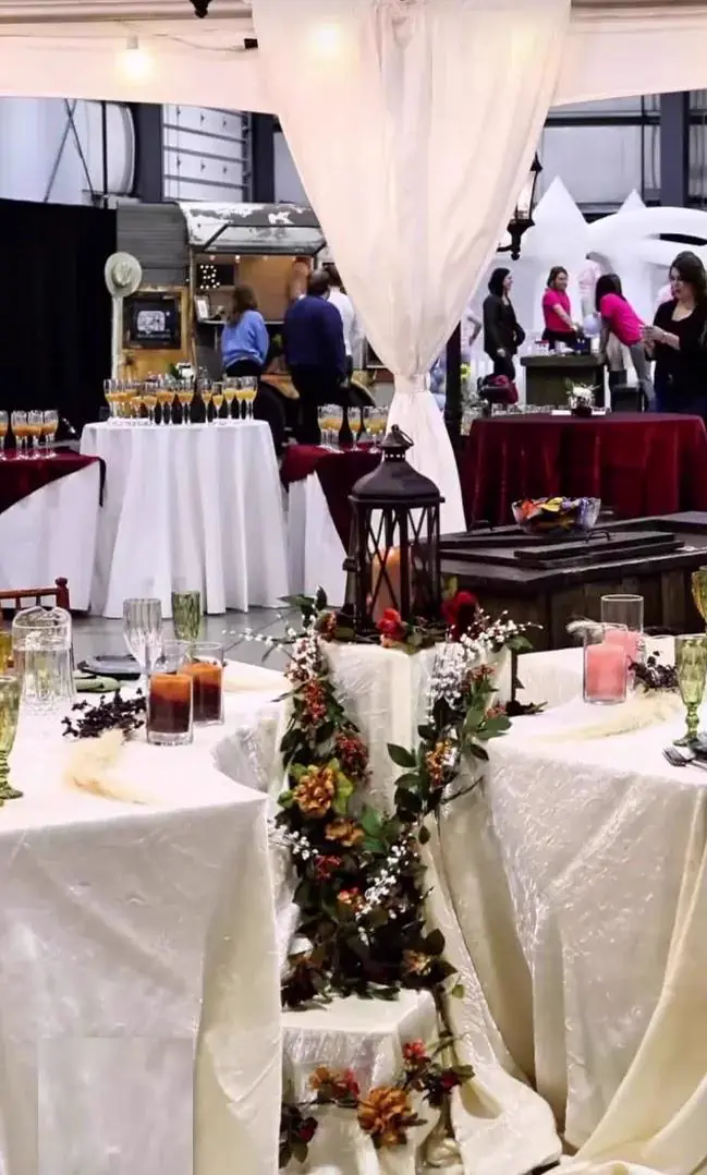 Display tables in white and burgundy linens with a black lantern and greenery at an indoor event Display tables in white and burgundy linens with a black lantern and greenery at an indoor event