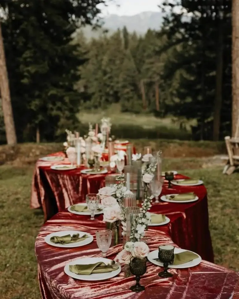 Long banquet table in a burgundy velvet cloth with green goblets and candles among pine trees Long banquet table in a burgundy velvet cloth with green goblets and candles among pine trees