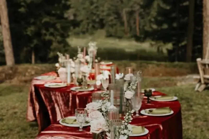 Long banquet table in a burgundy velvet cloth with white flowers and glassware among trees