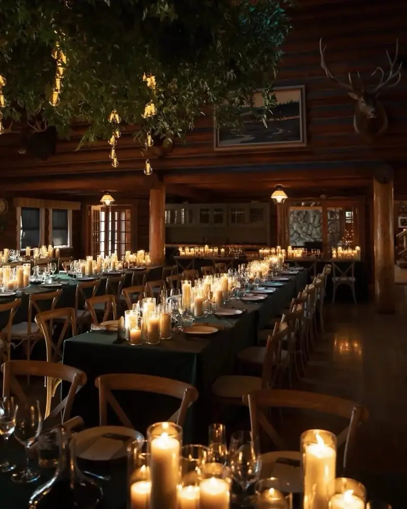 Long tables in dark green cloths lined with pillar candles in a log-walled room with hanging greenery