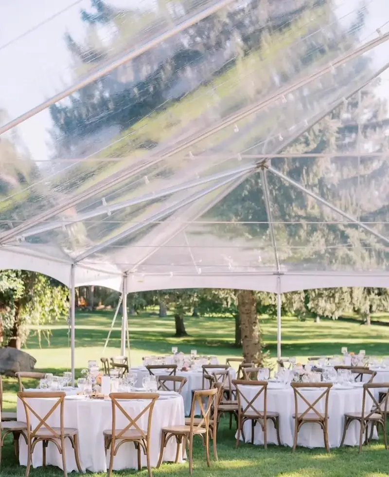 Round tables in white cloths with cross-back chairs and white flowers under a clear-top tent on a lawn Round tables in white cloths with cross-back chairs and white flowers under a clear-top tent on a lawn
