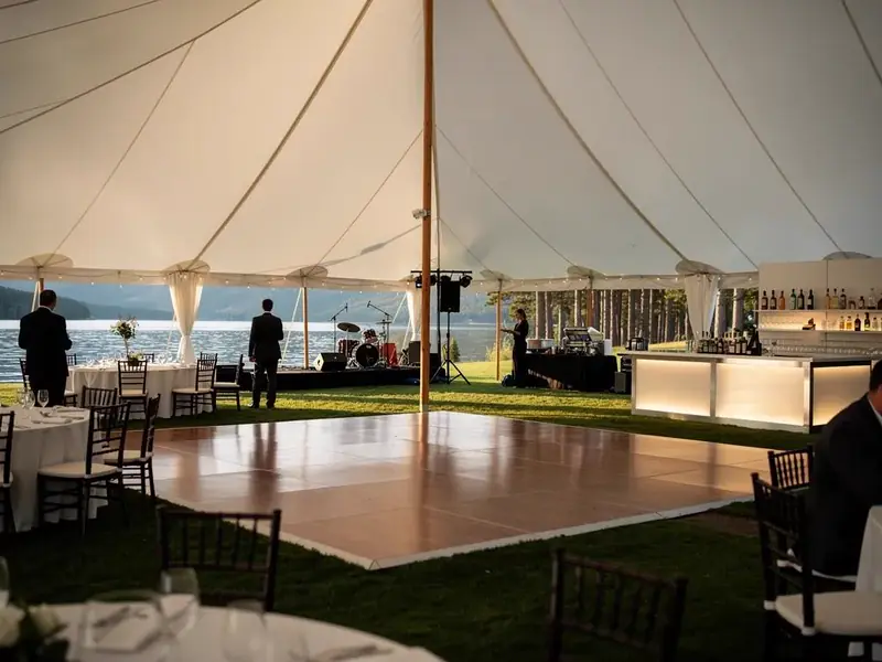 Wood dance floor, round tables and a lit bar under a white tent on a lawn beside a lake and mountains