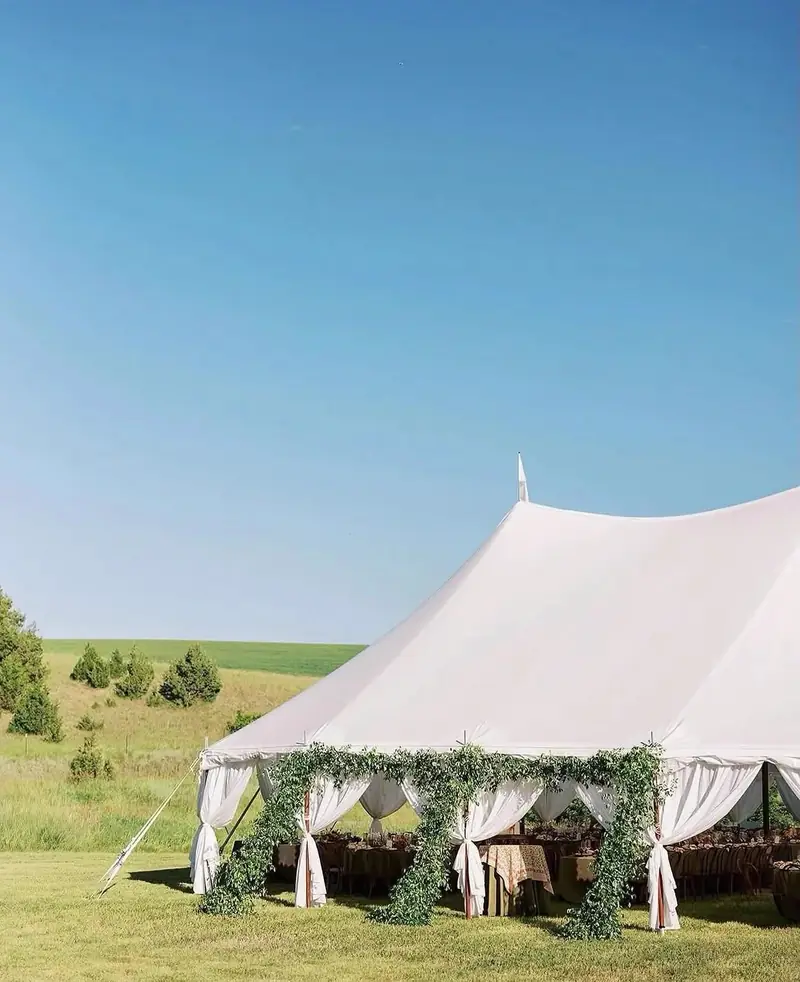 High-peak white tent with a greenery garland along the front on a field with green hills High-peak white tent with a greenery garland along the front on a field with green hills