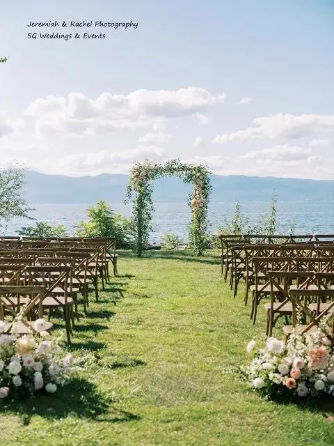 Rows of dark wood ceremony chairs facing a greenery arch on a lawn beside a lake