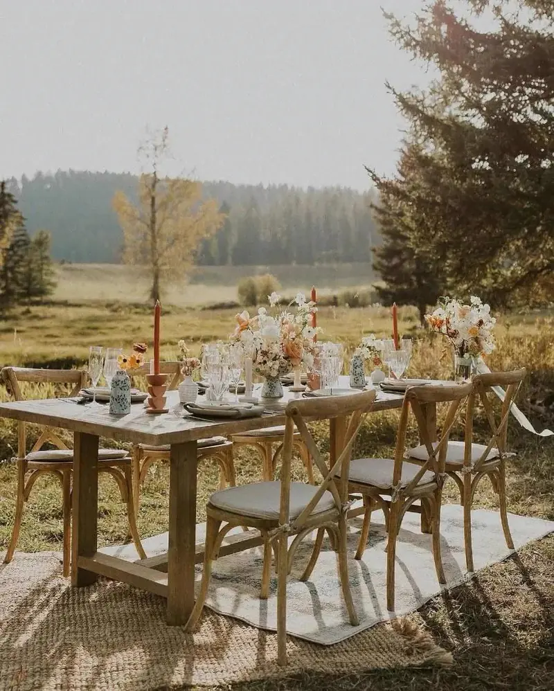 Long wood table with cross-back chairs and amber taper candles on a woven rug in a grassy meadow Long wood table with cross-back chairs and amber taper candles on a woven rug in a grassy meadow