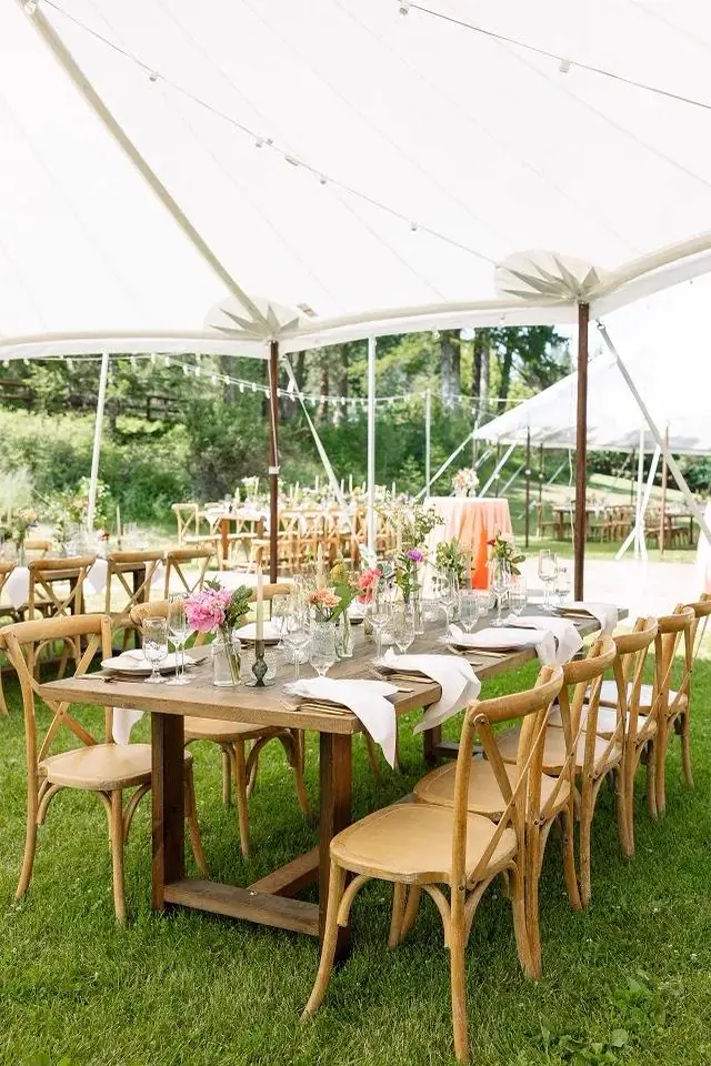Long wood table with cross-back chairs and coral flowers under an open-sided white tent on a lawn Long wood table with cross-back chairs and coral flowers under an open-sided white tent on a lawn