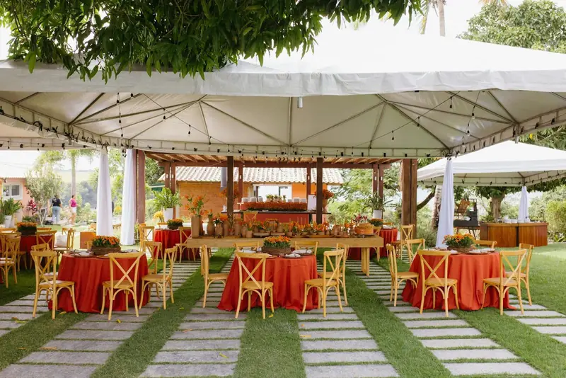 Round tables in orange-red cloths with cross-back chairs under a white canopy on a lawn Round tables in orange-red cloths with cross-back chairs under a white canopy on a lawn