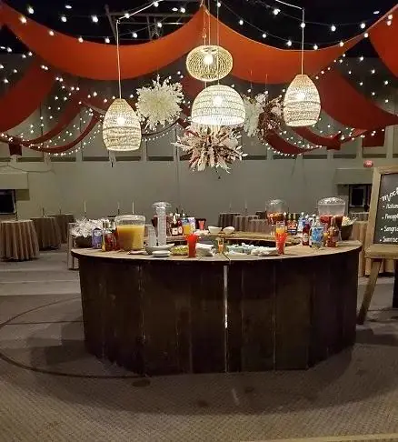 Curved wood bar with drink dispensers under rust draping and string lights beside a chalkboard menu