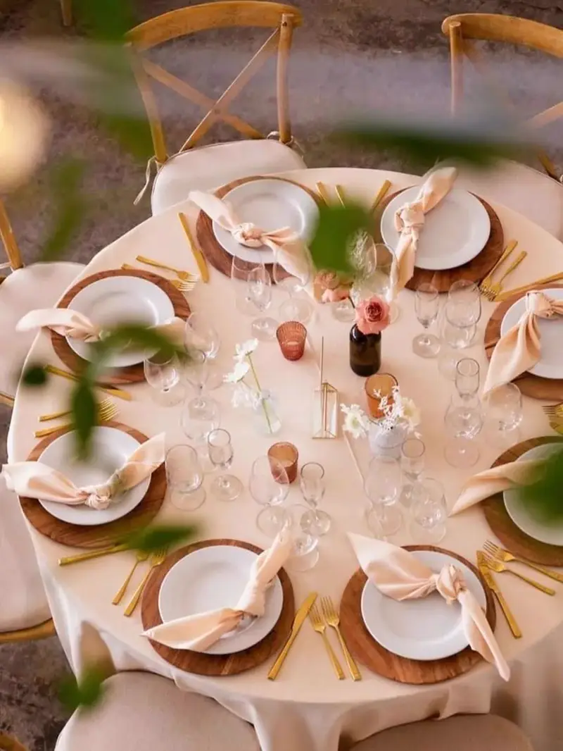 Overhead view of a round table with a cream cloth, wood chargers, blush napkins, candles and cross-back chairs