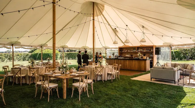 Sailcloth tent over a lawn with wood tables, cross-back chairs, a wood bar and a lounge area Sailcloth tent over a lawn with wood tables, cross-back chairs, a wood bar and a lounge area