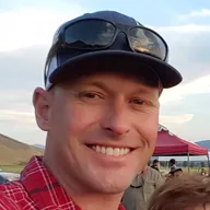 Scott Eastman, owner of Celebrate Event Rentals, smiling outdoors in a red plaid shirt and dark cap, with a red event tent and dry hills behind him