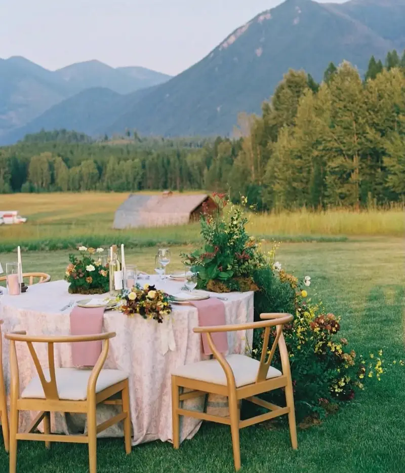 Table for two with a lace cloth, blush runner, taper candles and flowers on grass with mountains behind Table for two with a lace cloth, blush runner, taper candles and flowers on grass with mountains behind