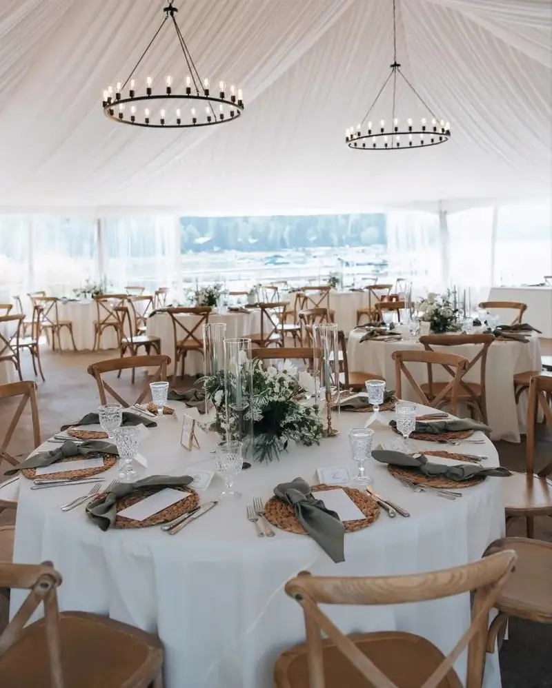 Round tables with white cloths, sage napkins and cross-back chairs under wagon-wheel chandeliers in a white tent