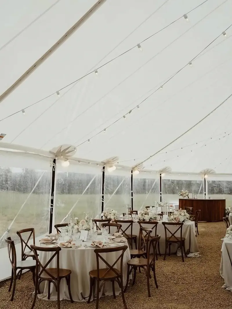 Inside a white tent with clear side walls, round tables in cream cloths, cross-back chairs and a wood bar Inside a white tent with clear side walls, round tables in cream cloths, cross-back chairs and a wood bar