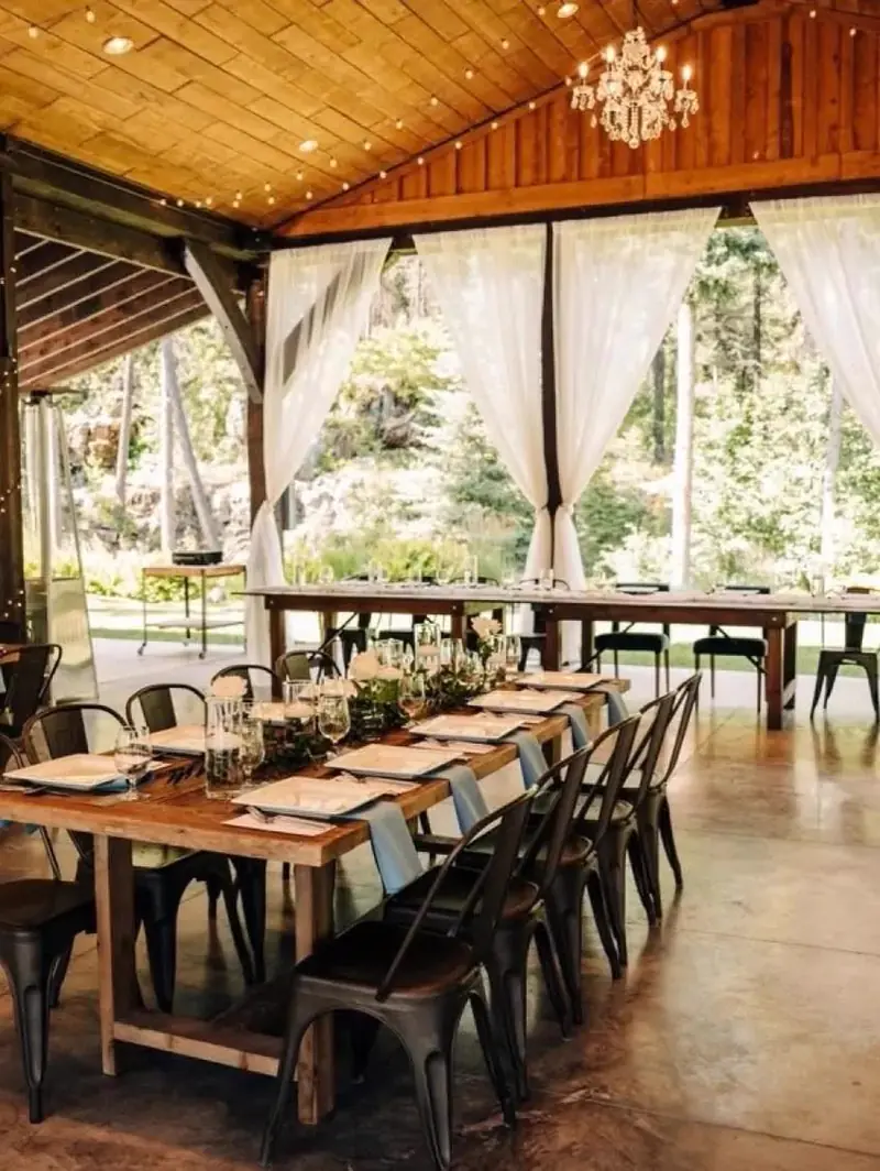 Long wood table with metal chairs under an open wood pavilion with sheer curtains and string lights Long wood table with metal chairs under an open wood pavilion with sheer curtains and string lights
