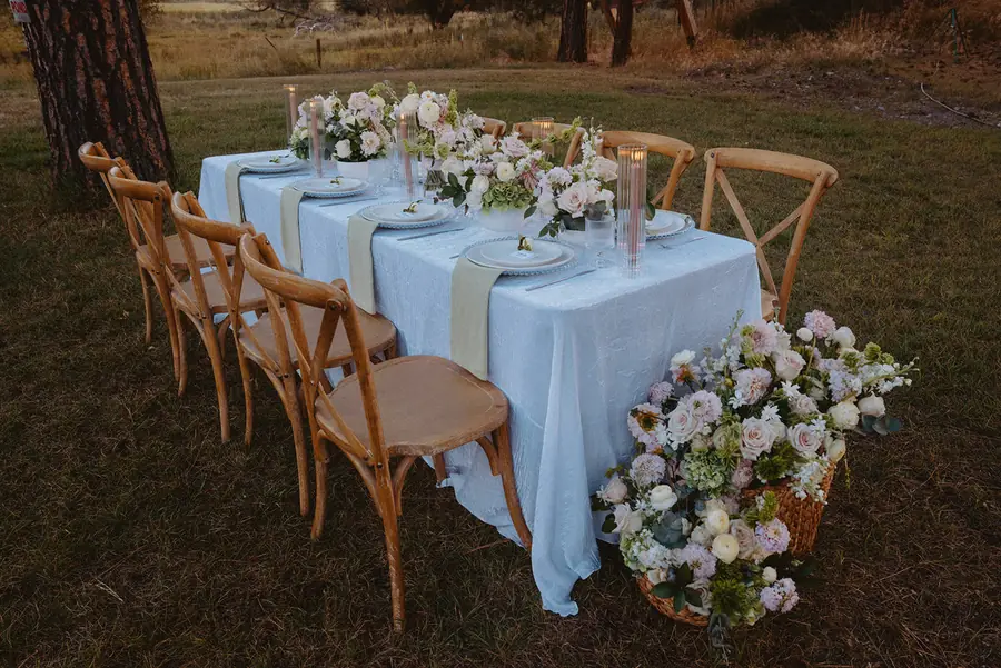 Long reception table with white linens and crossback chairs set under the pines