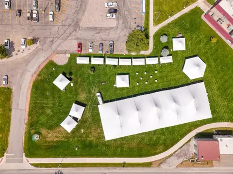 A large white event tent, a row of small booth tents and cocktail tables on a park lawn, seen from above
