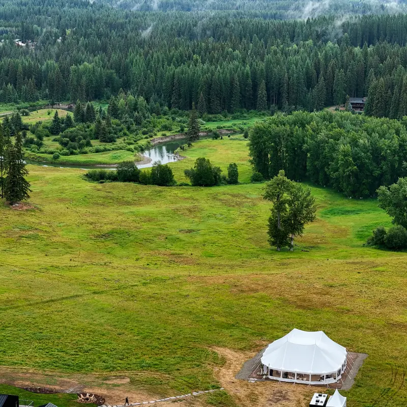 Aerial view of a white tent set in a wide green field near a winding river and dense forest