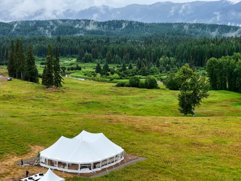Aerial view of a white event tent pitched in a wide green meadow below forested mountains Aerial view of a white event tent pitched in a wide green meadow below forested mountains