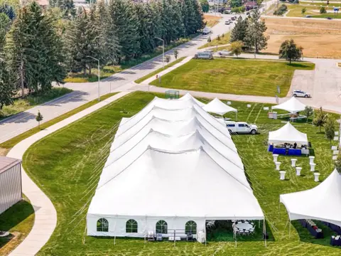 A long white tent with arched windows on a lawn beside a path and a row of trees