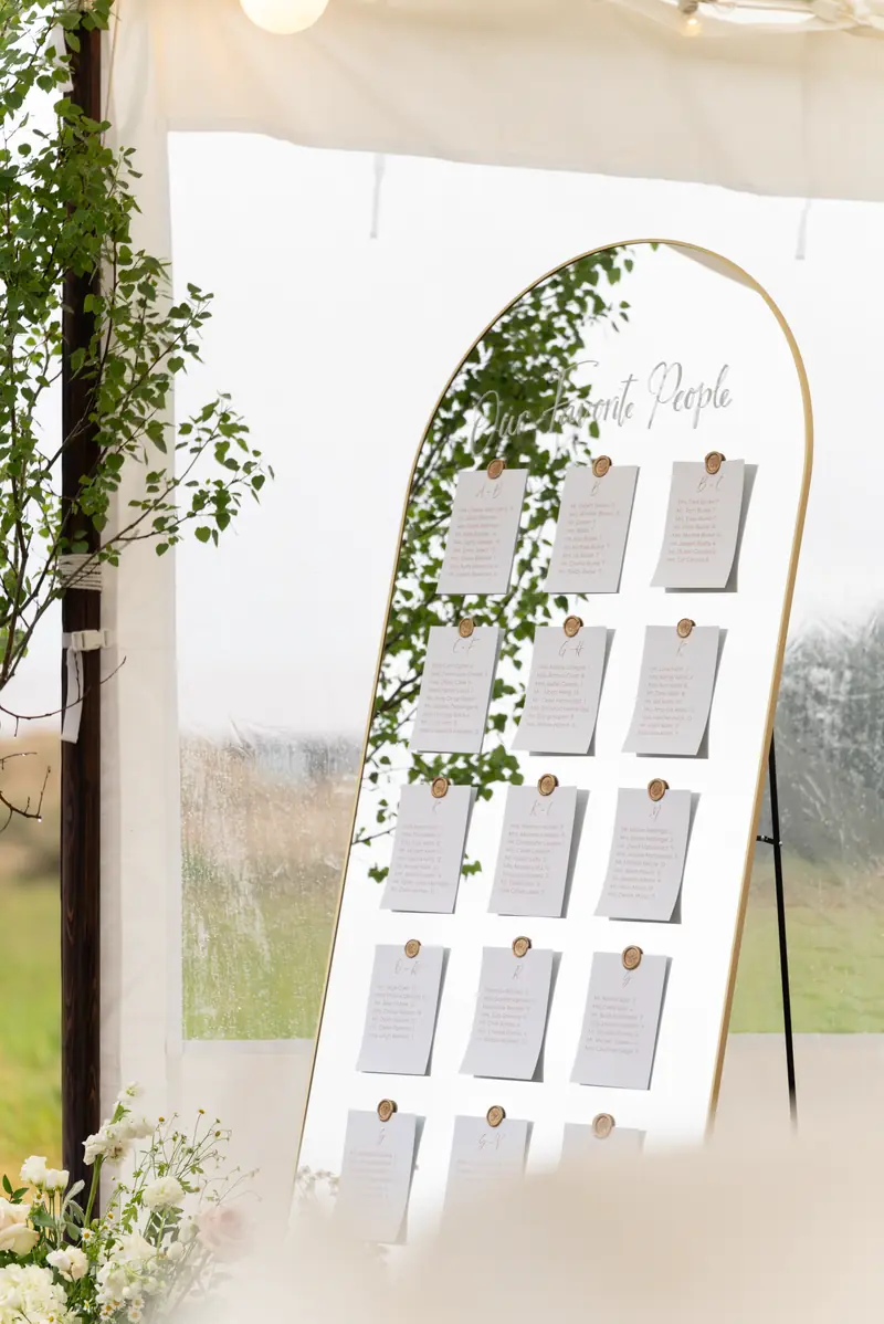 An arched seating chart board framed in greenery standing at the entrance of a tent