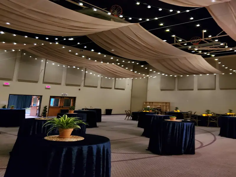 A wide conference hall with draped white ceiling fabric, string lights and cocktail tables set for guests