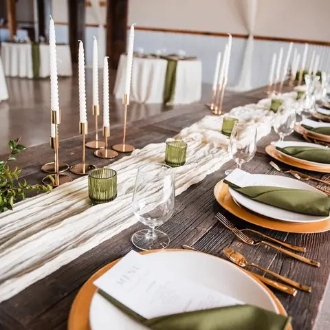 A cream table runner with an olive green napkin, white plates, a gold charger and brass candlesticks