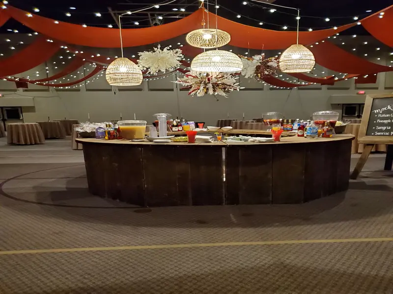 A curved wood beverage bar set under warm ceiling drapes and hanging lights in an event hall A curved wood beverage bar set under warm ceiling drapes and hanging lights in an event hall
