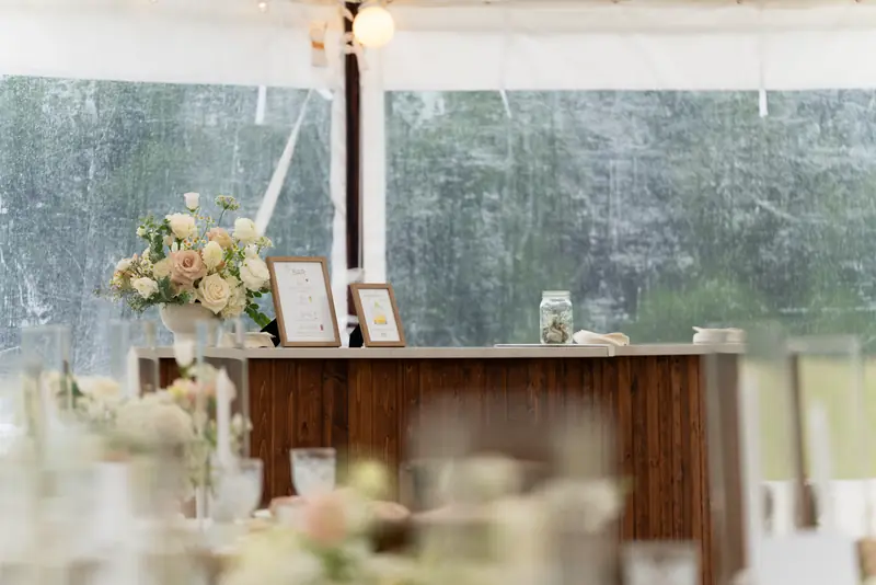 A dark wood bar under a white tent holding a floral arrangement, two framed signs and a glass jar A dark wood bar under a white tent holding a floral arrangement, two framed signs and a glass jar