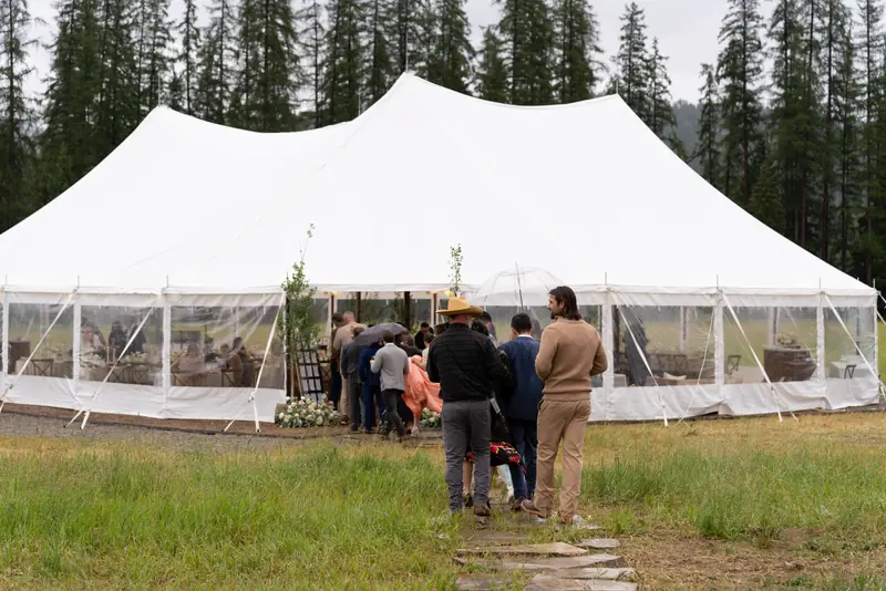 Guests gather at the open door of a white sailcloth tent set on a lawn below tall pines