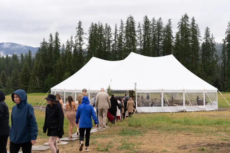 People walk along a stone path across a grassy field toward a large white event tent near tall pine trees