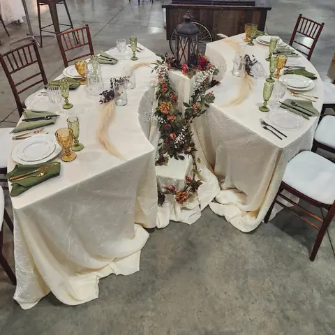 Two curved tables in ivory cloths with sage green napkins, white plates and amber and green glass goblets beside wood chairs