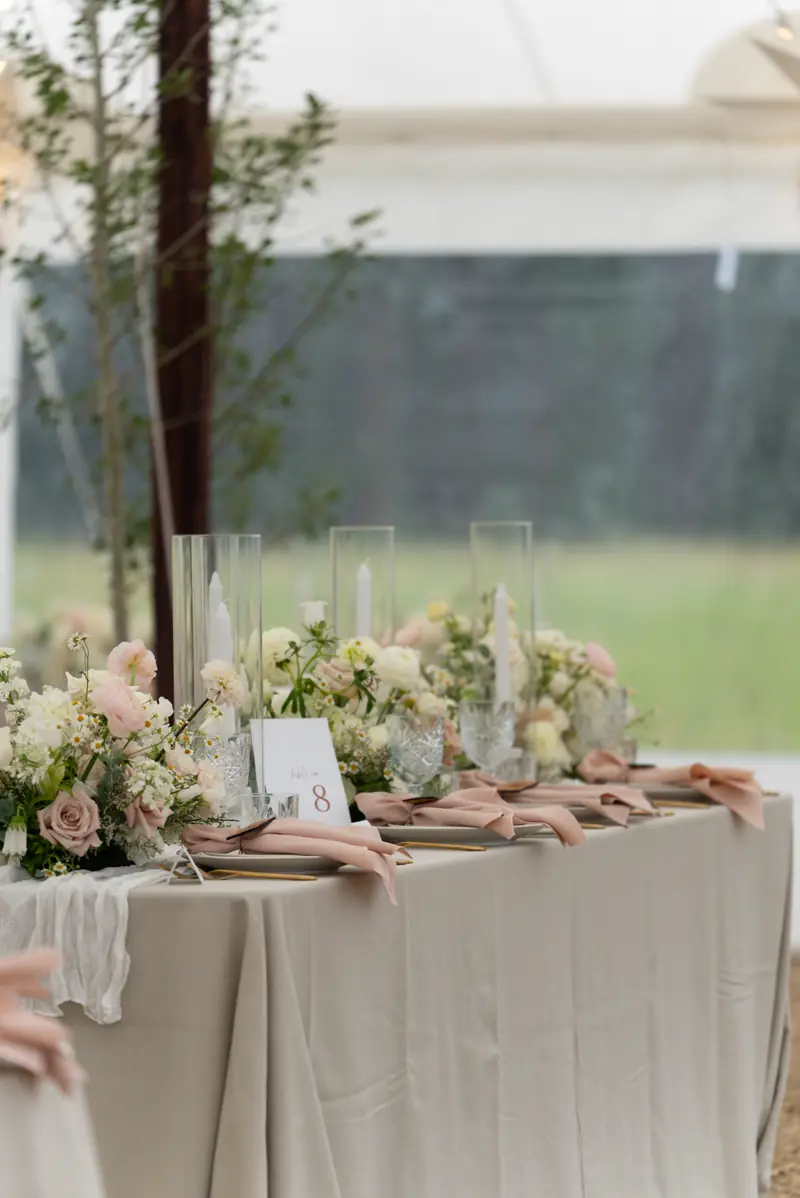 A long reception table in a beige tablecloth with pink flowers, tall glass candle cylinders and folded pink napkins
