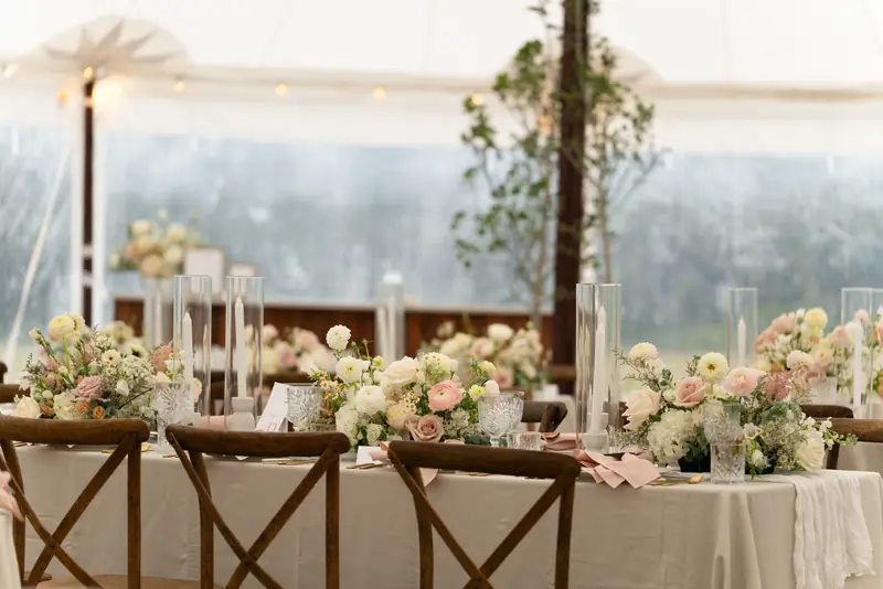 Wood chairs around a tent dining table dressed with pastel flowers, tall candle cylinders and glassware
