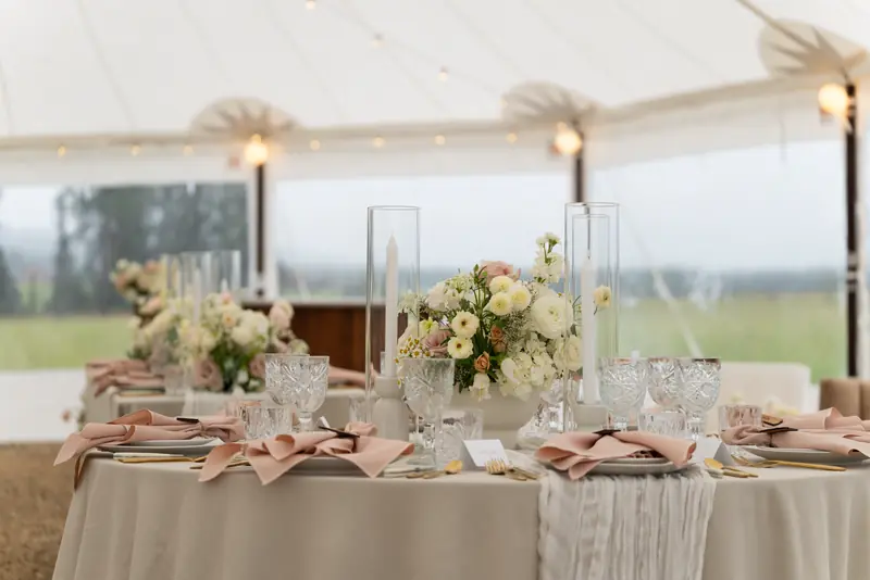 A round guest table in a beige cloth with a white gauze runner, blush napkins, candles and a pink and ivory centerpiece