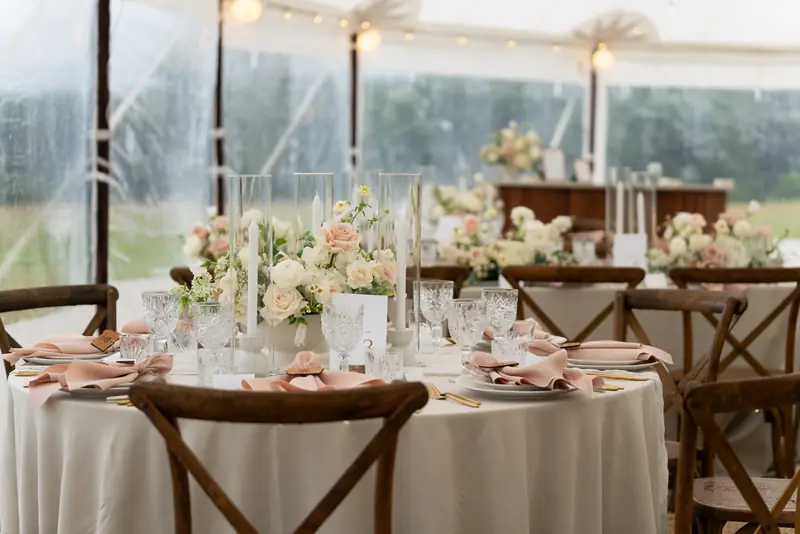 A dressed round table with wooden cross-back chairs, gold flatware, crystal glasses, candle cylinders and flowers, with clear tent walls behind