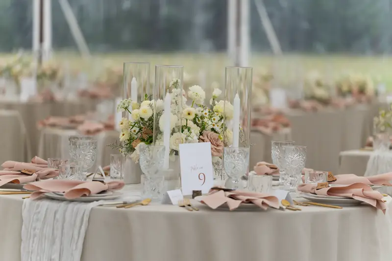 A round table set with blush pink napkins, gold flatware, crystal glasses, white flowers and a table number card