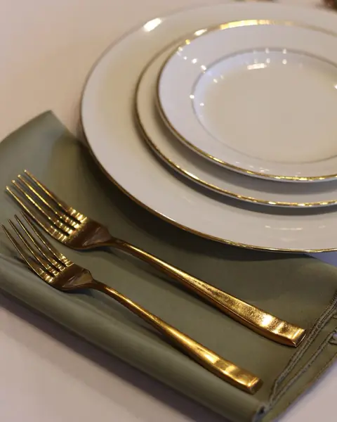 A sage green napkin with two gold forks beside stacked gold rimmed white plates on an off-white cloth