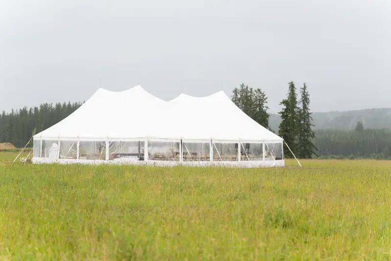 A white sailcloth tent with clear sidewalls in a tall grass field, with pine trees off to one side
