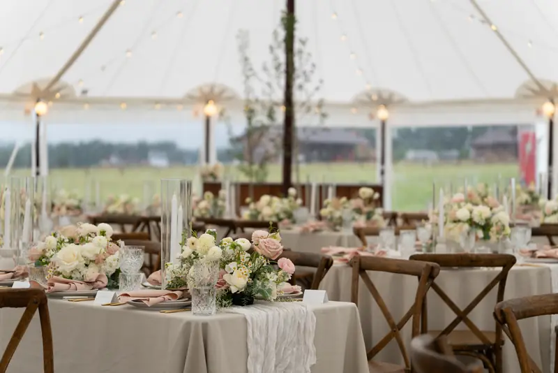Tables set with beige tablecloths, floral centerpieces, tall glass candles and wooden cross-back chairs inside an illuminated tent Tables set with beige tablecloths, floral centerpieces, tall glass candles and wooden cross-back chairs inside an illuminated tent