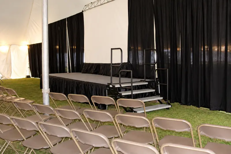A low stage with a black skirt and metal stairs at one end of a tent, facing rows of folding chairs A low stage with a black skirt and metal stairs at one end of a tent, facing rows of folding chairs