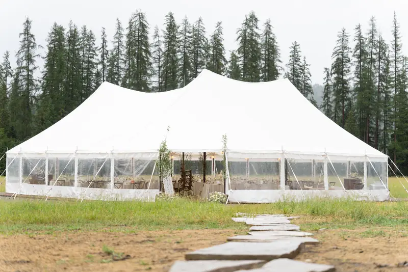 A stone path crosses a green lawn into a white sailcloth tent with tall dark pines behind it