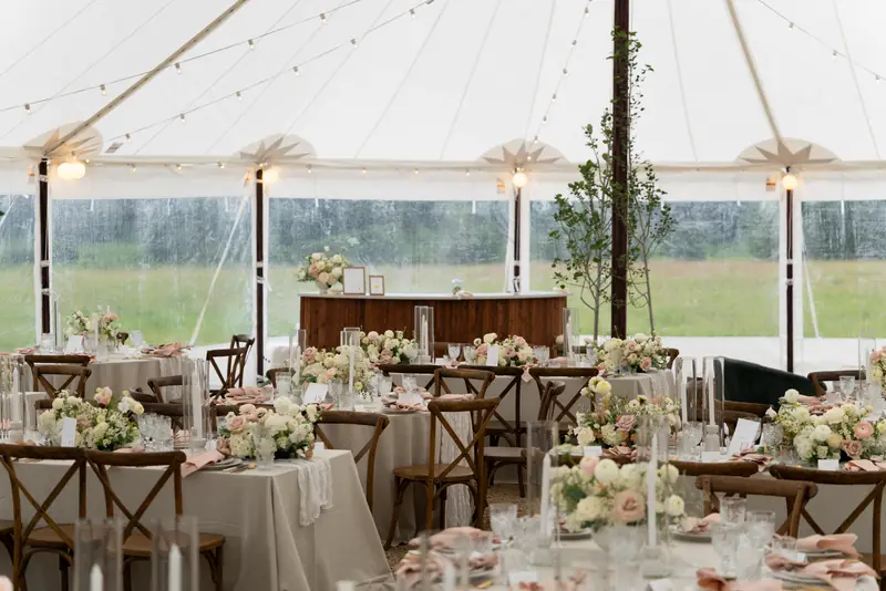 Tables with floral centerpieces and cross-back chairs inside a white tent with string lights and clear walls