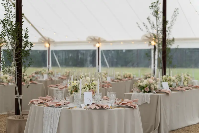 Decorated tables with beige tablecloths, floral centerpieces, crystal glasses and blush napkins inside a large white tent Decorated tables with beige tablecloths, floral centerpieces, crystal glasses and blush napkins inside a large white tent