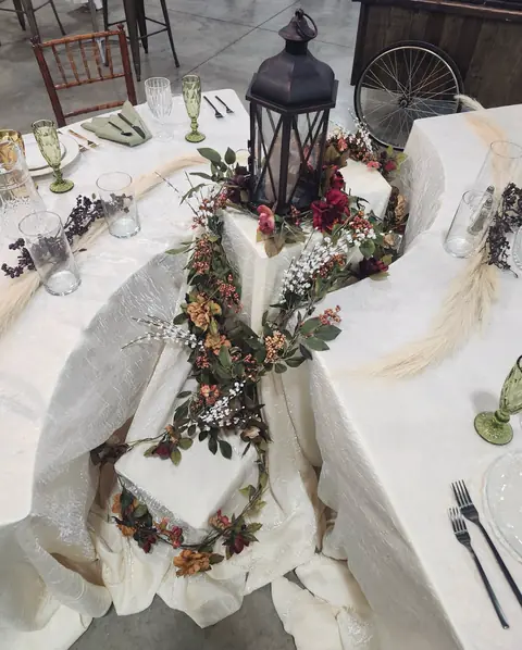 A tiered display table in draped cream fabric with a dark lantern, a floral garland, green goblets and sage napkins