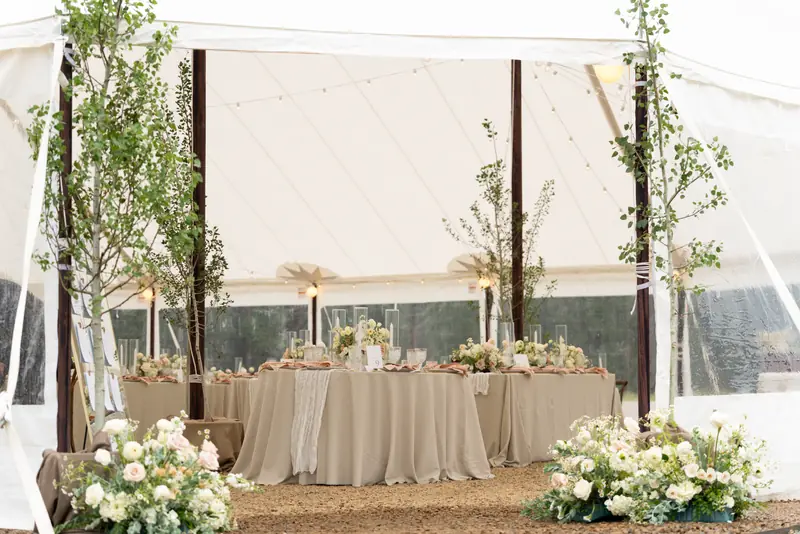 Two white floral arrangements on gravel outside a white tent filled with set tables and small trees