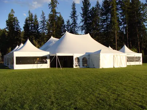 A white peaked tent with clear and arched window walls on a green lawn in front of tall pine trees