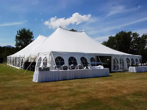 A white pole tent with arched window walls on a dry lawn, with long catering tables set outside under a blue sky