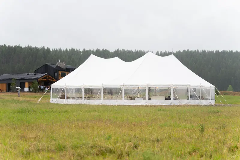 A white sailcloth tent with tall peaks and clear sidewalls in a grass field, tables set inside, with a dark wood lodge and pines behind it